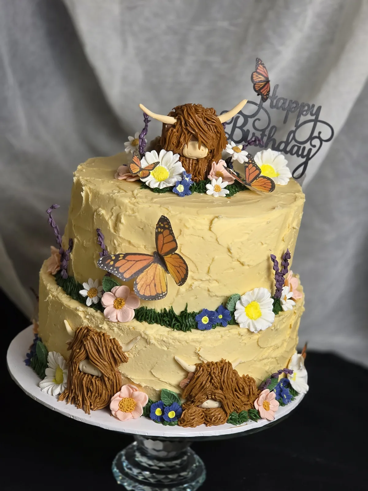 Two tier tan buttercream cake topped with a shaggy brown highland cow head, surrounded by colorful buttercream flowers and orange monarch butterfly decorations.