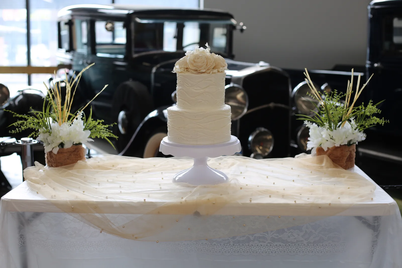 Two-tier white buttercream wedding cake with wave-textured icing and cream sugar roses on a white pedestal, flanked by floral arrangements in front of vintage cars.