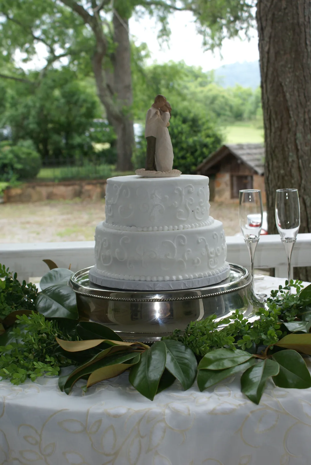 Two tier white wedding cake with piped swirl patterns and a romantic couple figurine topper displayed on a silver pedestal surrounded by fresh greenery.