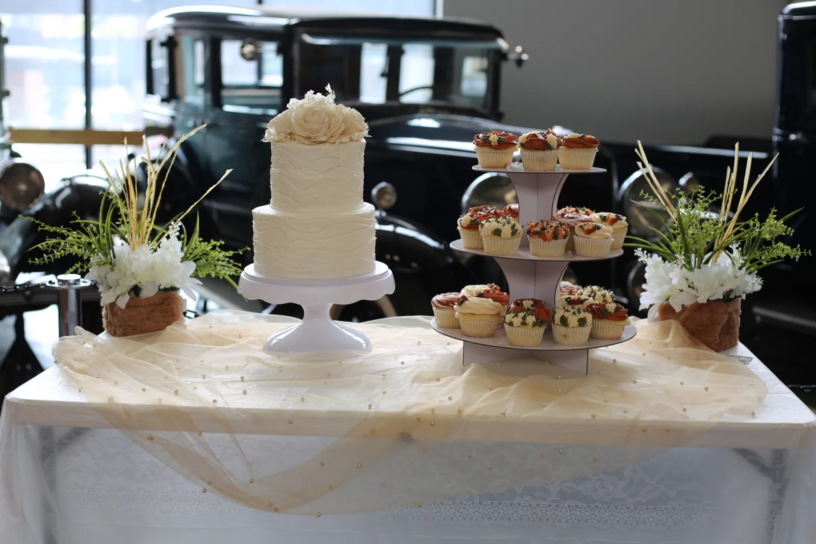 Two-tier white buttercream wedding cake with wave-textured icing and a sugar flower topper beside a three-tier stand of rose cupcakes, set in front of vintage cars.