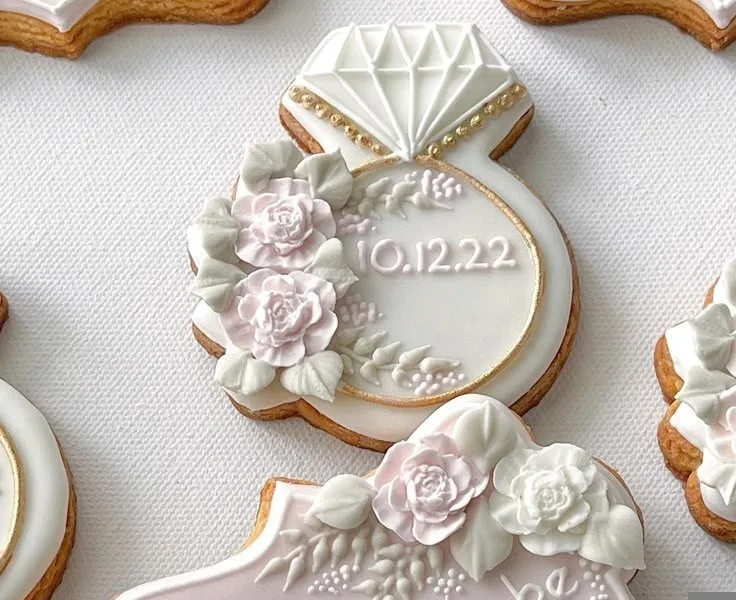 Wedding cookies iced in white with a diamond ring, the date 10.12.22 and piped white flowers.