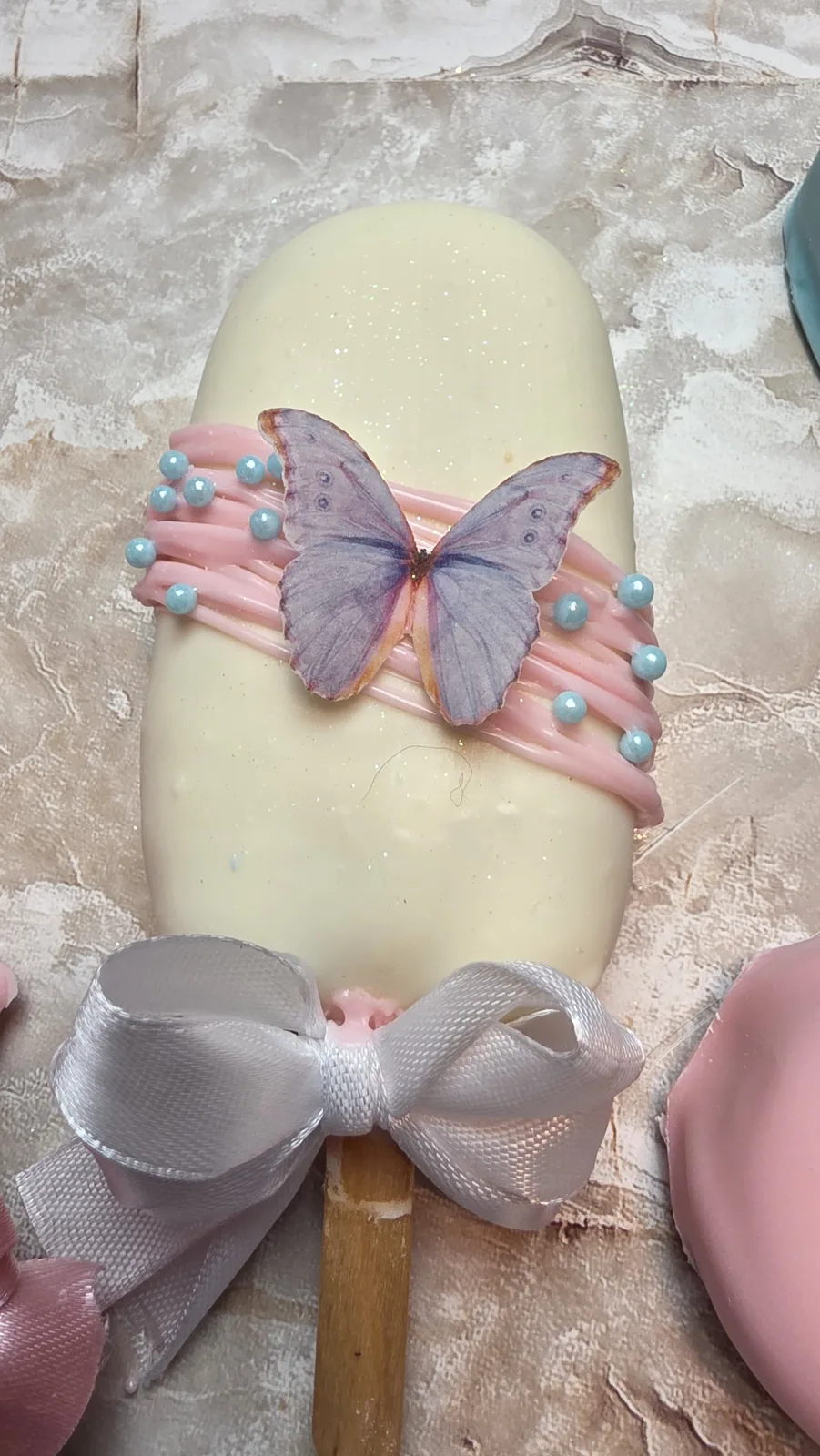 White chocolate coated cakesicle decorated with pink drizzle, blue sugar pearls, and an edible purple butterfly image, tied with a silver ribbon.