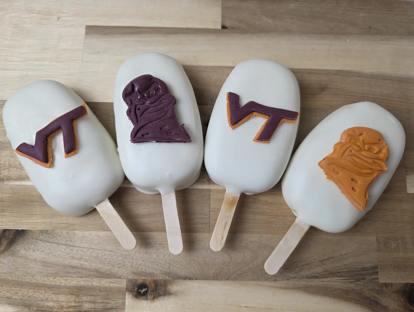 Four white chocolate coated cakesicles on wooden sticks featuring purple and orange decorative shapes including character bust designs and stylized logos.