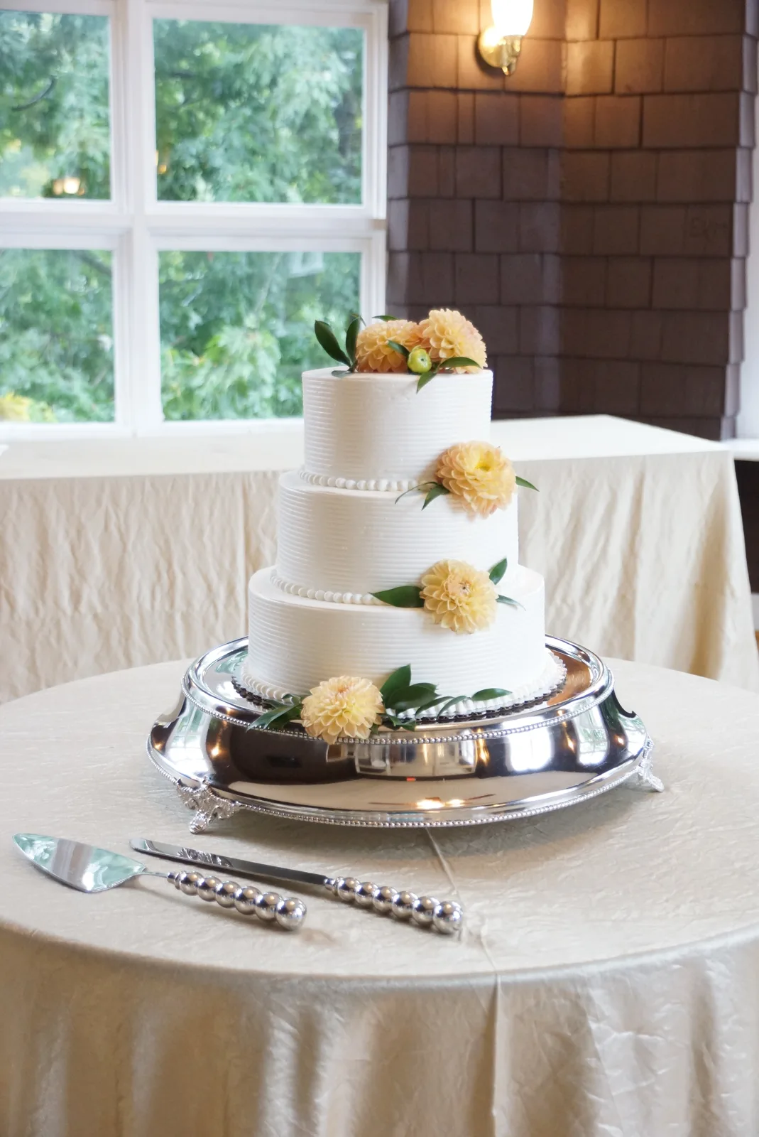 Three tiered white textured buttercream wedding cake adorned with peach yellow dahlias and green leaves on a polished silver stand.