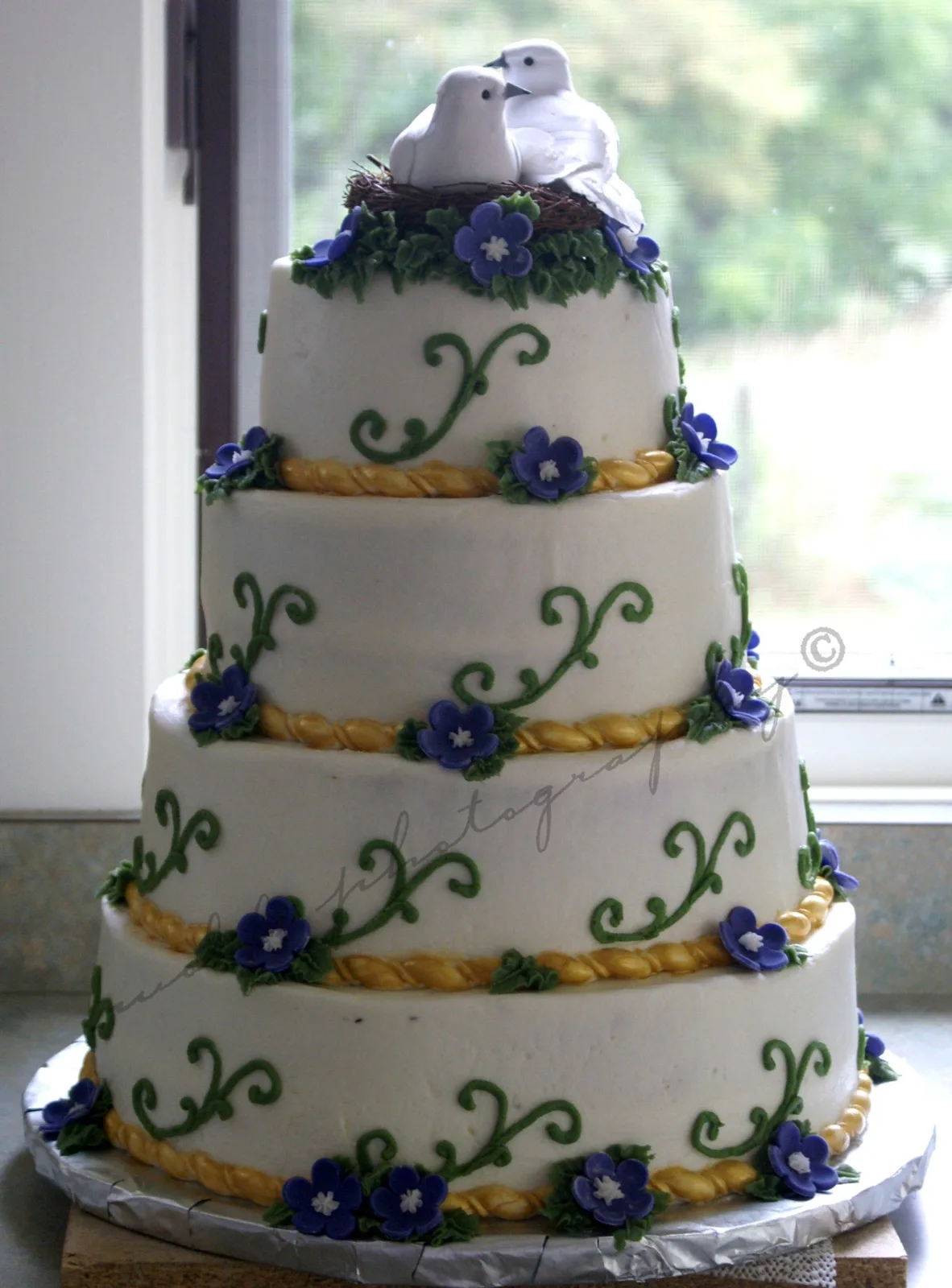 Four tiered white wedding cake topped with two white dove figurines in a nest, decorated with purple flowers, green vine scrollwork, and gold braided piping between tiers.