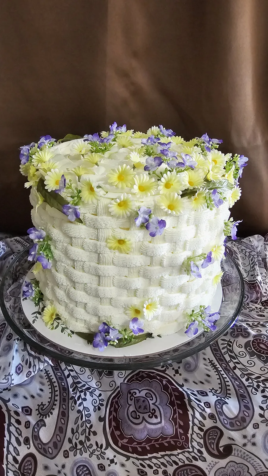 A tall white frosted cake with a basketweave textured design, topped with white roses, yellow daisies, and purple blooms cascading down the sides.