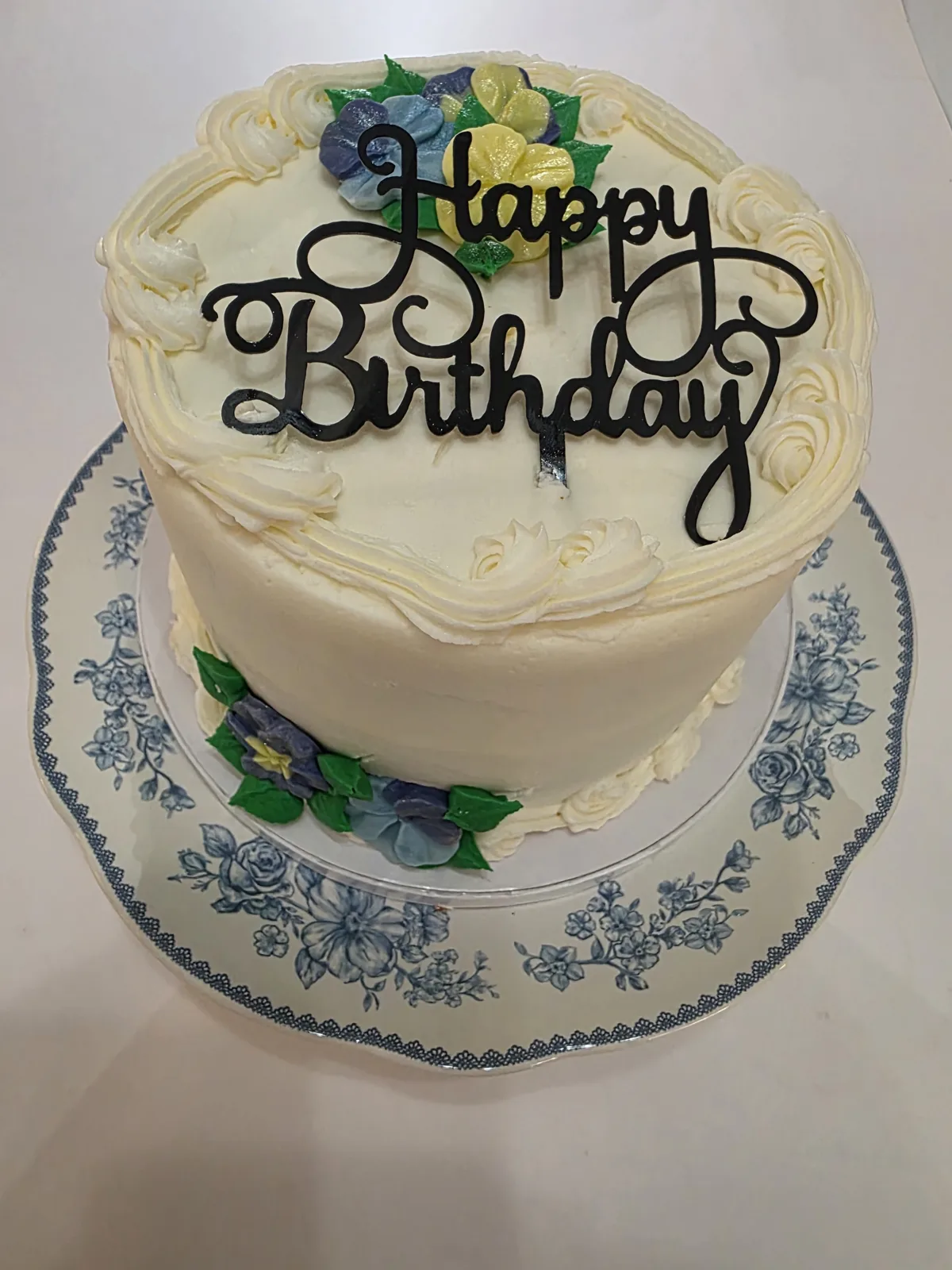 Tall single tier white frosted birthday cake decorated with buttercream rosettes, blue purple and yellow flowers with green leaves, and a black script topper.