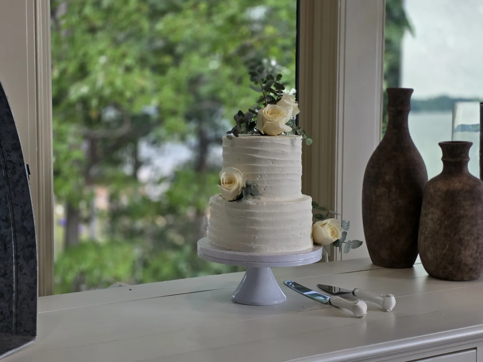 Two tier white textured buttercream wedding cake decorated with ivory roses and eucalyptus leaves on a white pedestal stand.