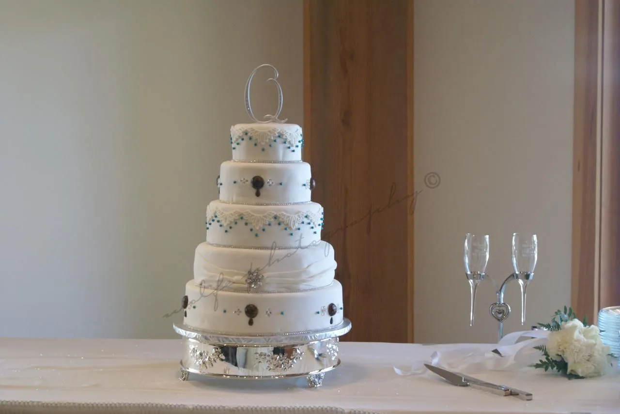 Four tier white fondant wedding cake with lace detailing, teal dot accents, brown jewel embellishments, silver beading, and a silver letter Q topper on a silver stand.