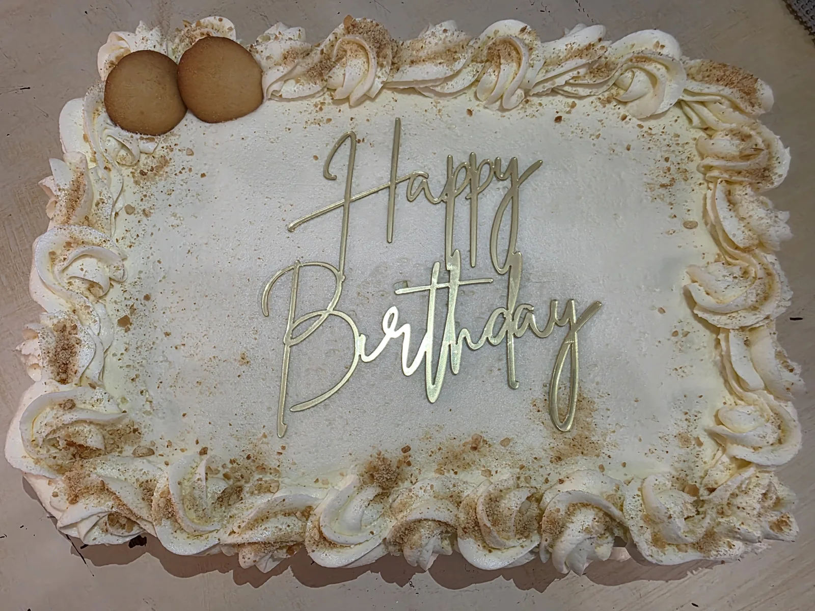 Rectangular white frosted cake with piped shell borders, gold Happy Birthday script, crumb dusting, and two vanilla wafers on top corner.