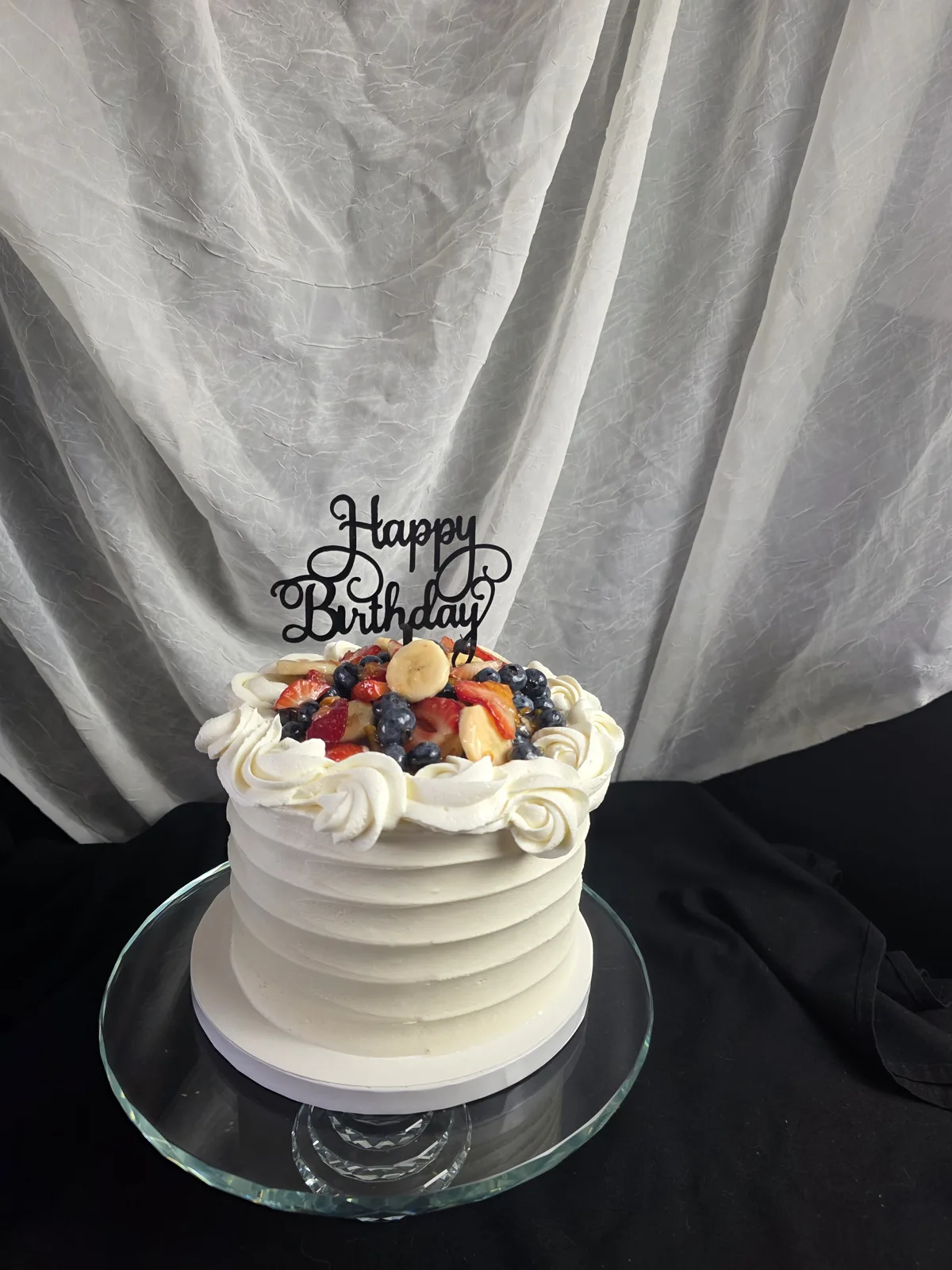 Small white frosted cake with ribbed buttercream sides, topped with strawberry, blueberry, and banana pieces surrounded by white frosting rosettes.