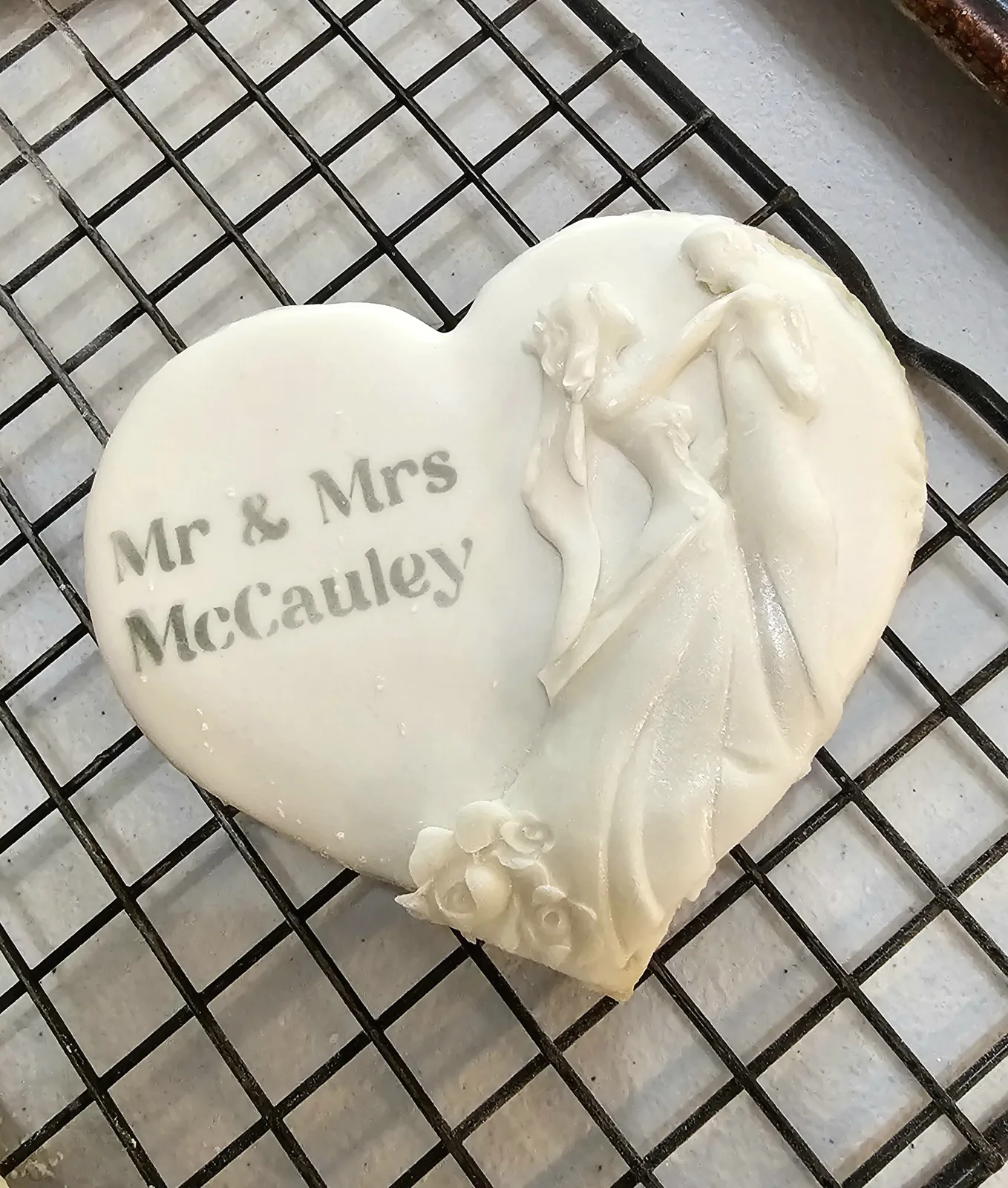 Heart shaped white iced sugar cookie decorated with draped fondant and piped floral clusters accenting stamped text on a wire rack.