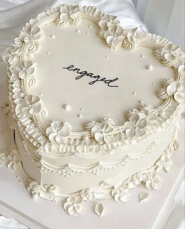 Elegant ivory heart shaped cake adorned with piped buttercream florals, ruffles, and pearl dots around the borders.