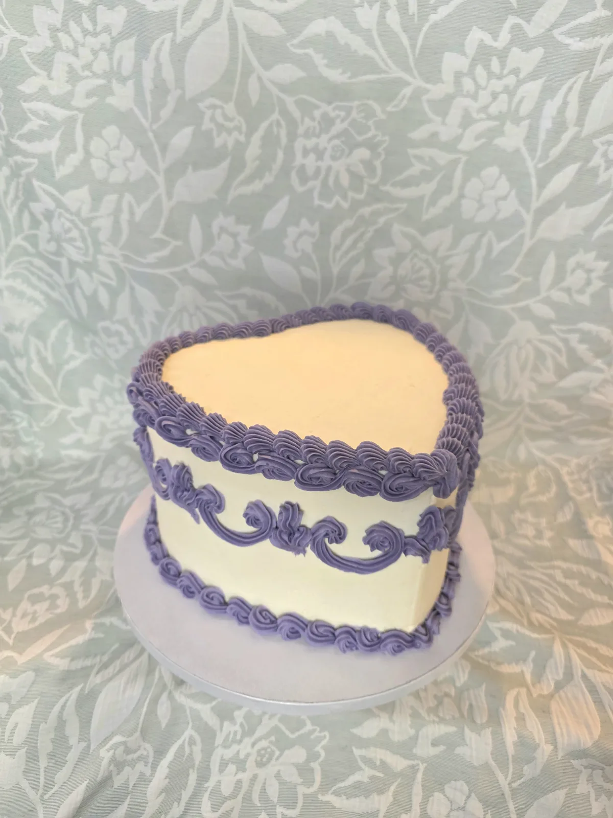 White heart shaped cake decorated with intricate lavender purple piped frosting borders, swags, and scrollwork on a silver cake board.