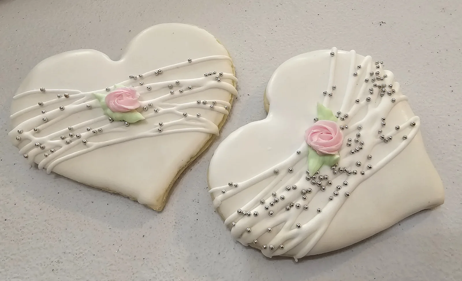 Two heart shaped sugar cookies covered in smooth white icing with delicate white icing drapes, silver dragées, and a pink royal icing rose with green leaves at the center.