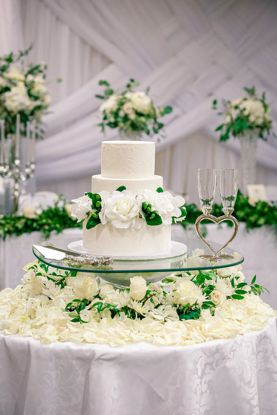 Two tier white wedding cake with textured lace patterned fondant, decorated with white roses and green leaves, displayed on a glass stand with silver server and heart shaped toasting flutes.