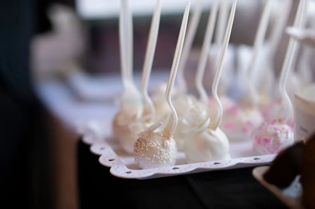 White chocolate cake pops with pearl sprinkles and pink speckles on a scalloped white tray.