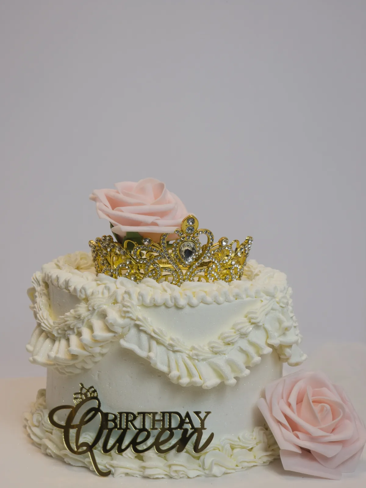 White buttercream cake with ruffled frosting layers, topped with a gold jeweled tiara, a pink rose, and a gold Birthday Queen sign.