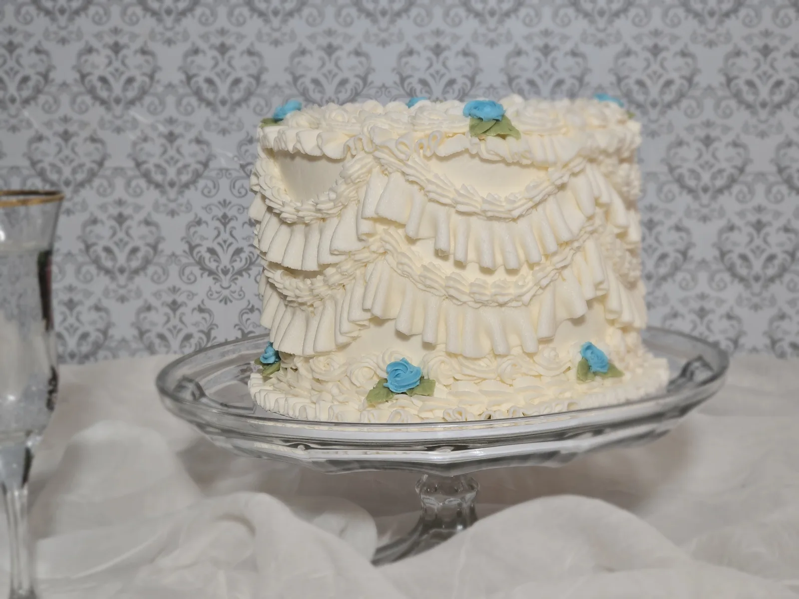White frosted cake featuring layered ruffle design and small blue rosebuds with green leaves, displayed on a clear glass pedestal stand.