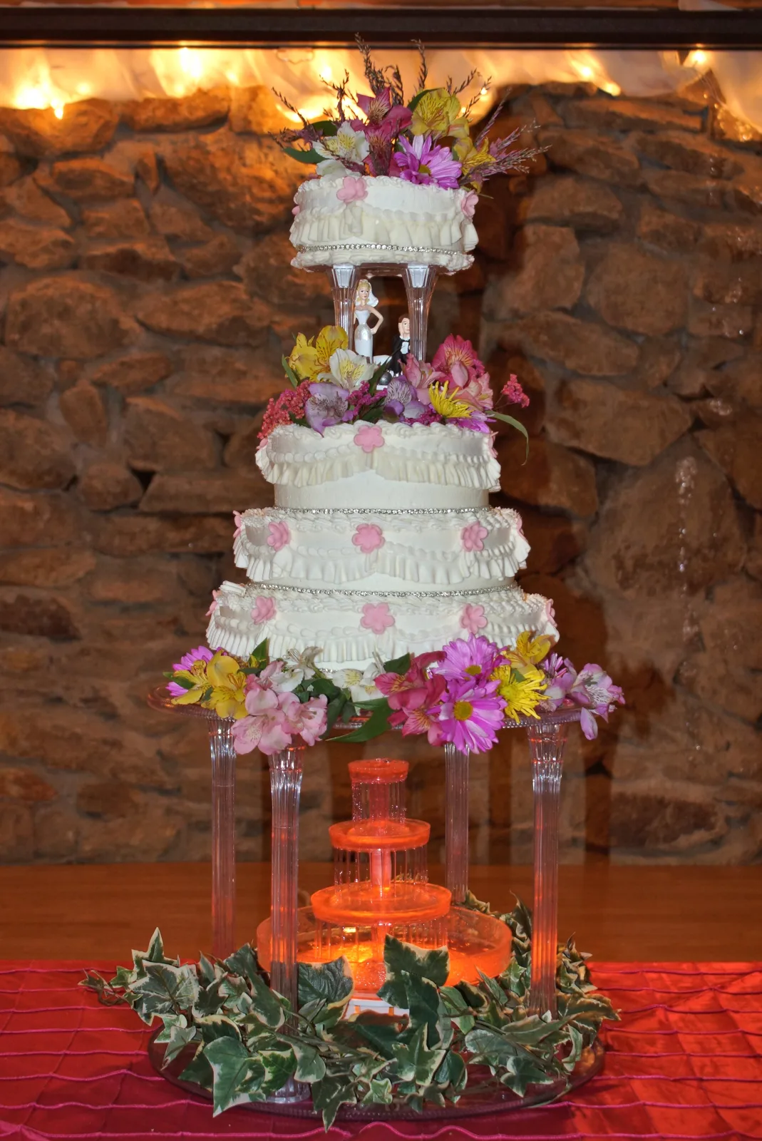 Four tiered white wedding cake with ruffled fondant and pink sugar flowers, surrounded by fresh purple, pink, and yellow blooms above a lit orange fountain.