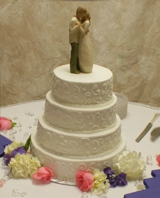 Three tiered white wedding cake decorated with piped scrollwork patterns, topped with an embracing bride and groom figurine, surrounded by pink roses and white hydrangeas.