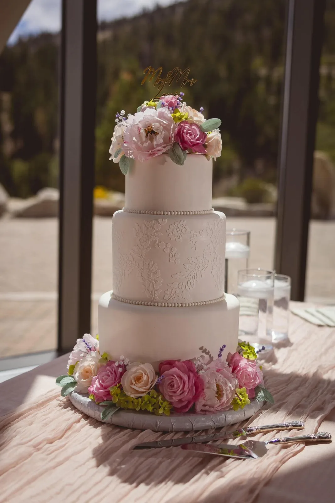 Three tier white wedding cake with lace textured middle tier, topped with pink and cream sugar flowers and a gold Mr and Mrs topper, with matching blooms at the base.