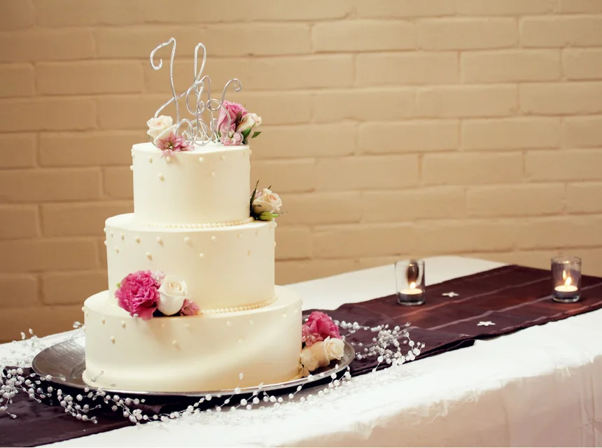 Three tier smooth white fondant wedding cake decorated with small pearl dots, pink carnations, ivory roses, and a silver wire monogram topper on a silver stand.