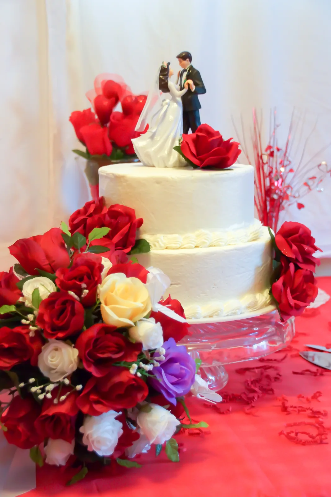 Two tier white frosted wedding cake topped with a bride and groom figurine and adorned with red white cream and purple roses on a red tablecloth.