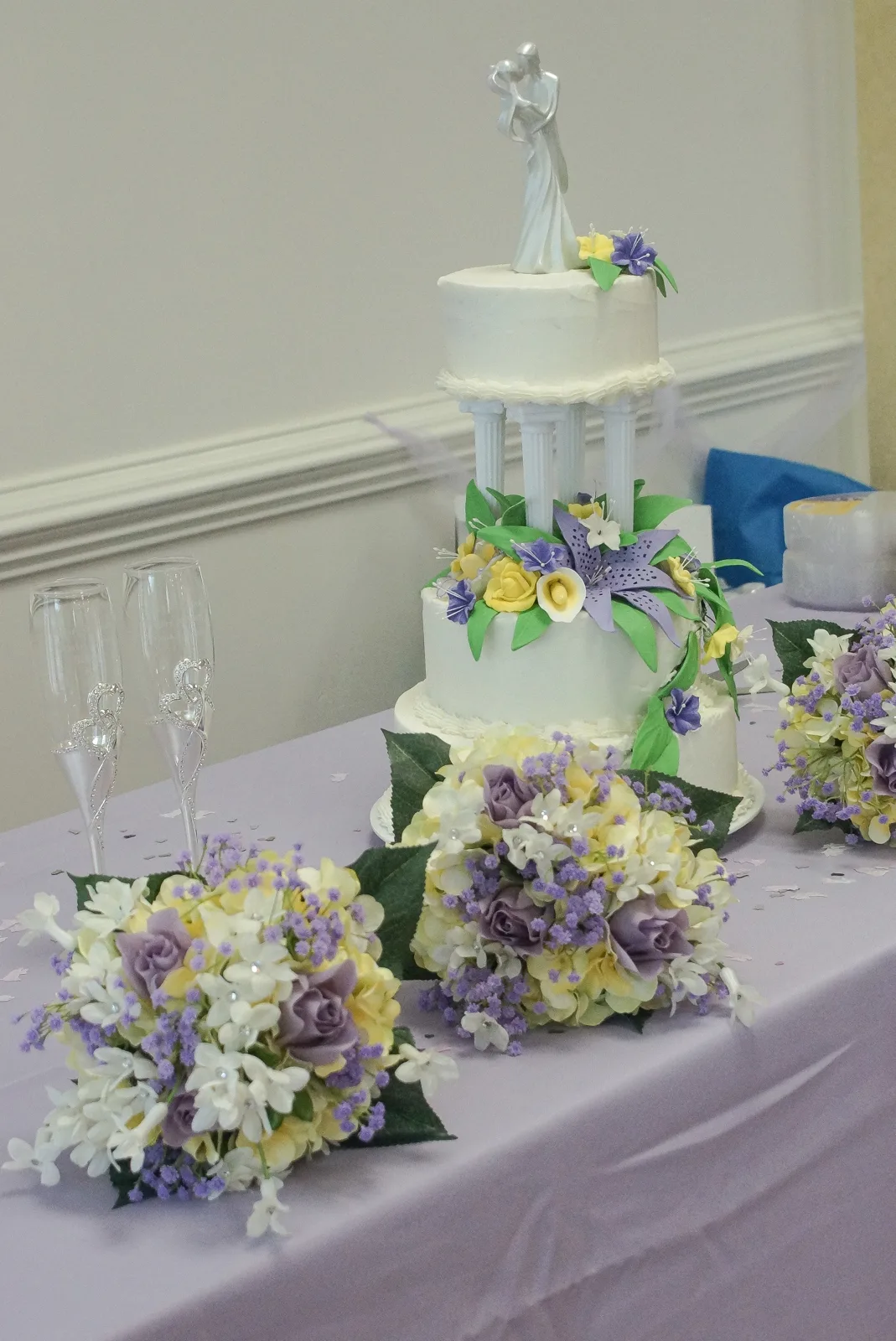 Two tiered white wedding cake separated by pillars, topped with a bride and groom figurine and decorated with yellow roses, purple lilies, and green leaves.