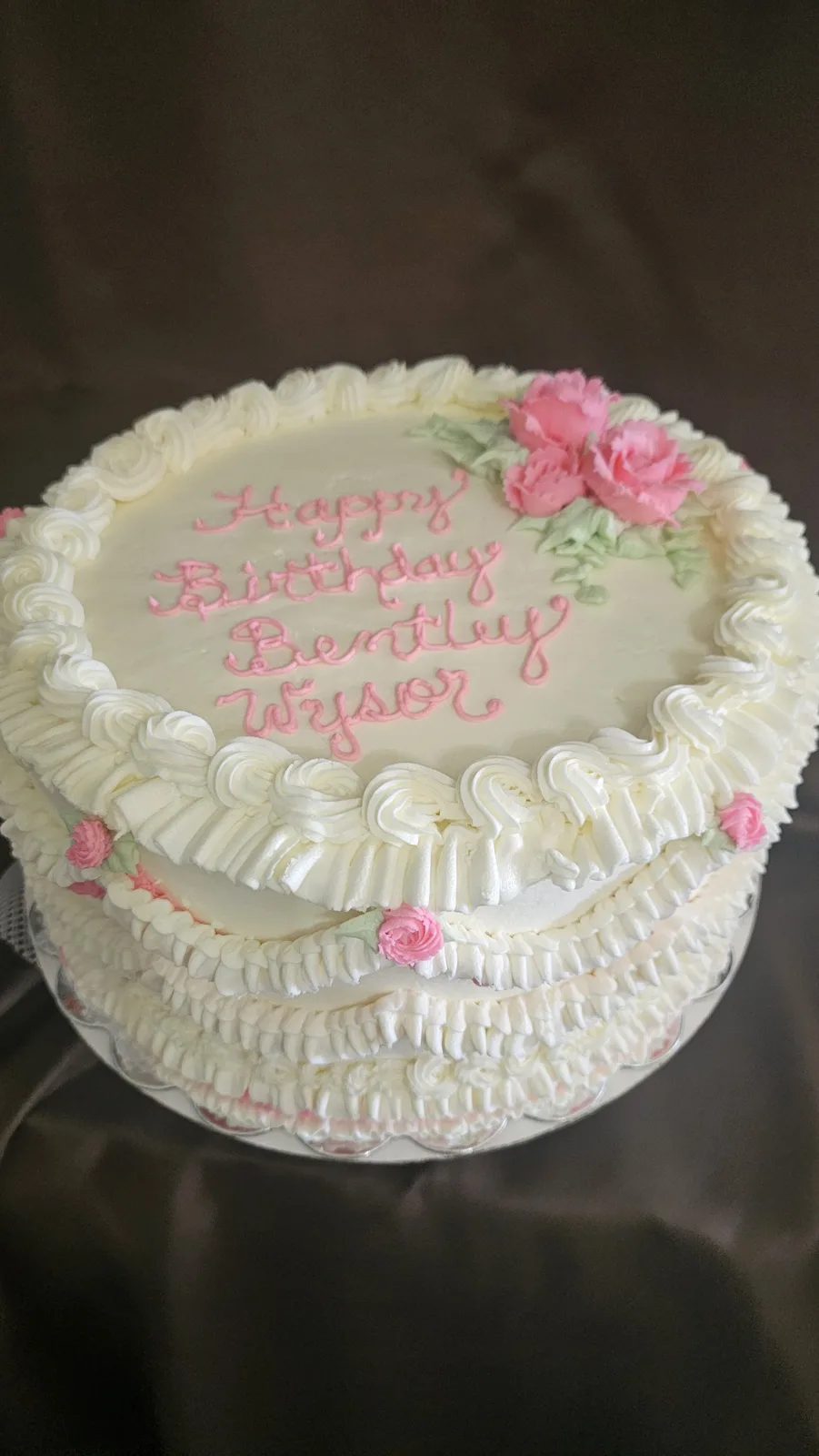 A tiered white frosted birthday cake decorated with pink piped script lettering, pink roses with green leaves, and ruffled white frosting borders on a silver cake board.