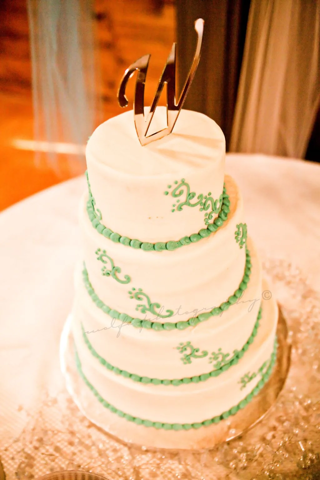 Three tiered white wedding cake decorated with green beaded borders, green scrollwork piping, and a tall gold metallic monogram topper.