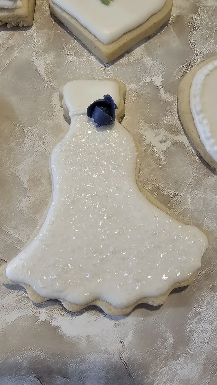 A sugar cookie shaped like a wedding dress covered in smooth white icing with a small navy blue fondant rose at the neckline, displayed on lace fabric.