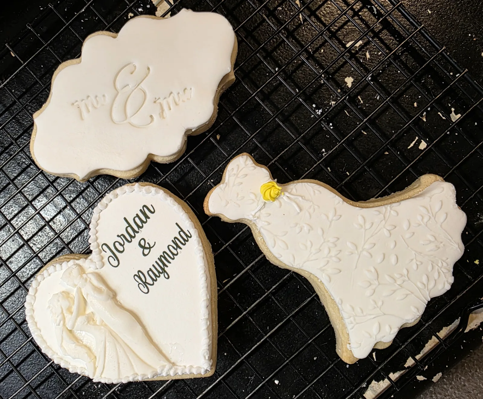 Three white iced wedding cookies on a black rack including a plaque embossed Mr and Mrs, a heart with a bride and groom silhouette, and a dress with leaf details.