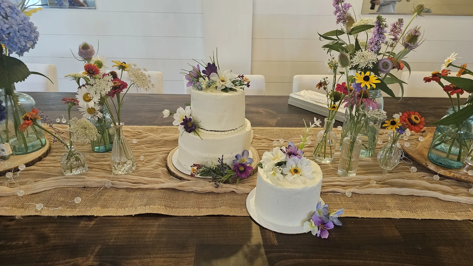 Two tier and single tier white buttercream wedding cakes decorated with fresh purple, white, and yellow flowers on wood slice bases.