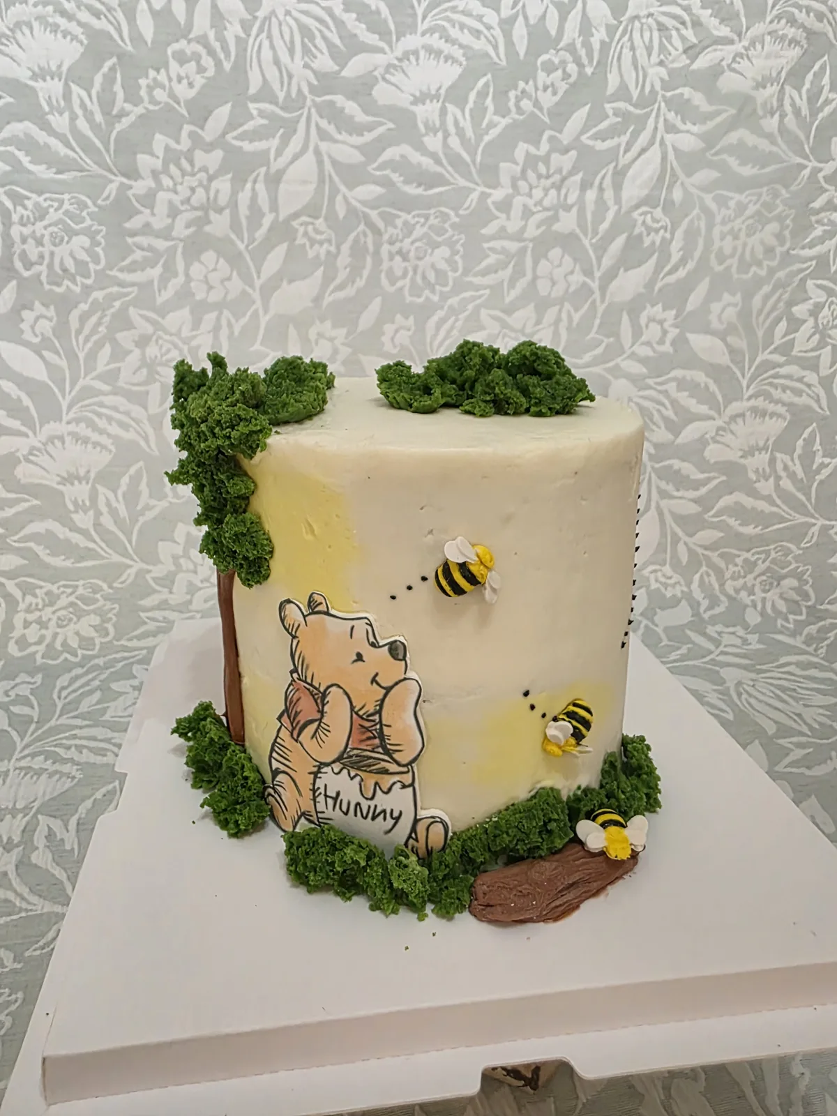 Tall cream frosted cake decorated with piped green foliage, brown tree branches, a hand painted Winnie the Pooh holding a hunny pot, and three small yellow and black bee figures.