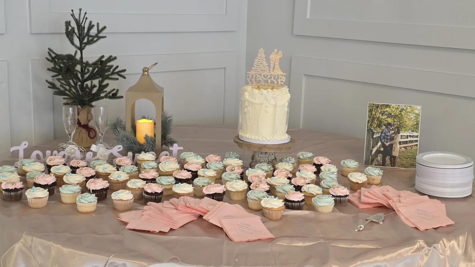A blush dessert table featuring a small white frosted cake with a gold couple and tree topper, surrounded by rows of pink, white, and mint cupcakes with pearl accents.