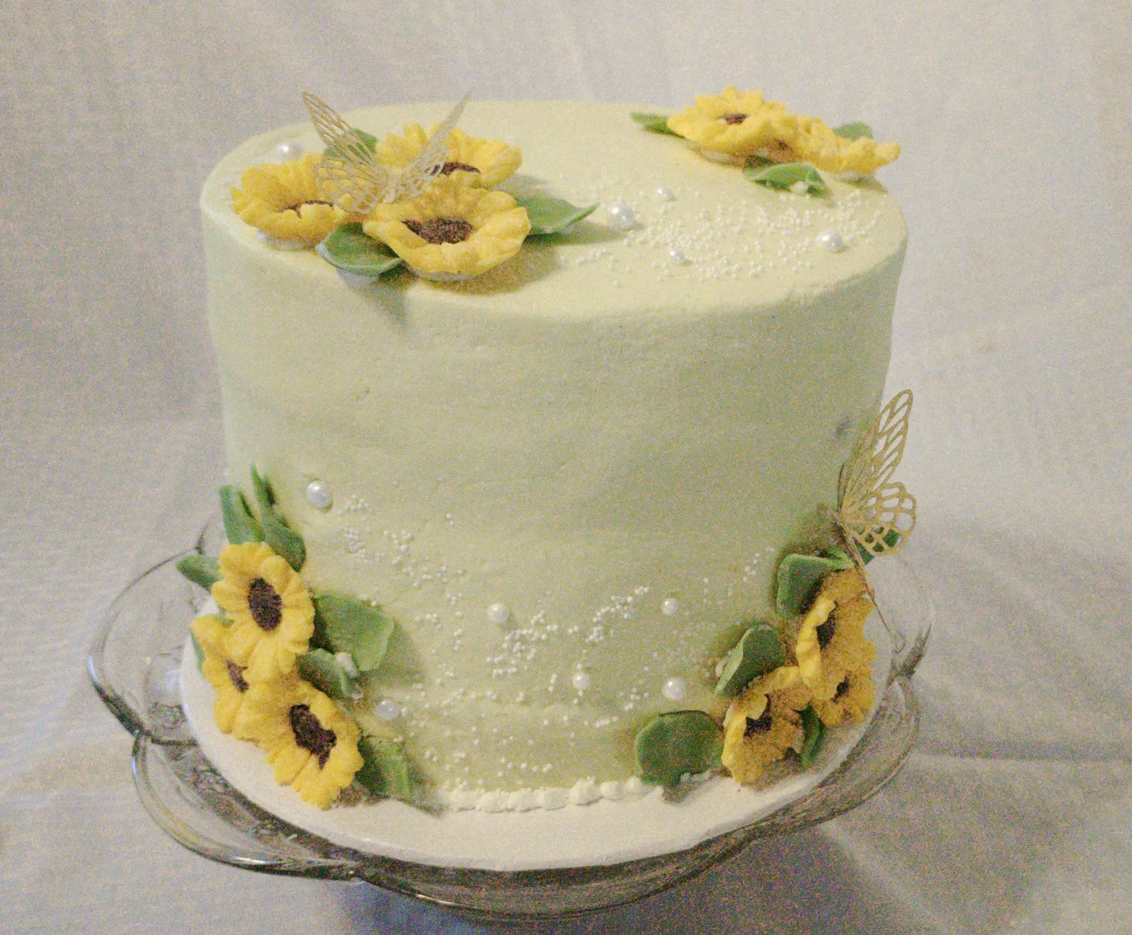 Tall ivory buttercream cake adorned with yellow piped flowers, green leaves, gold butterfly accents, and scattered white sugar pearls on a glass stand.