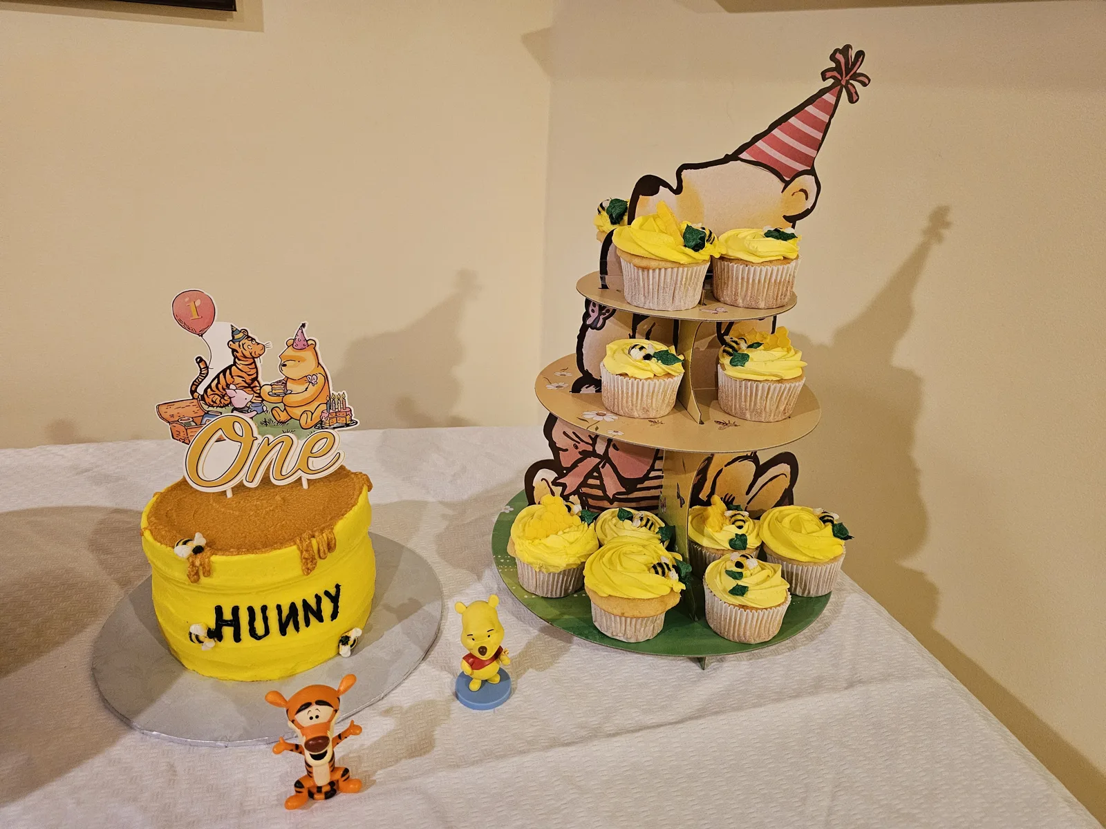 A yellow frosted cake designed like a honey pot with "HUNNY" text and a Winnie the Pooh themed topper reading "One" beside a tiered stand of matching cupcakes with bee decorations.