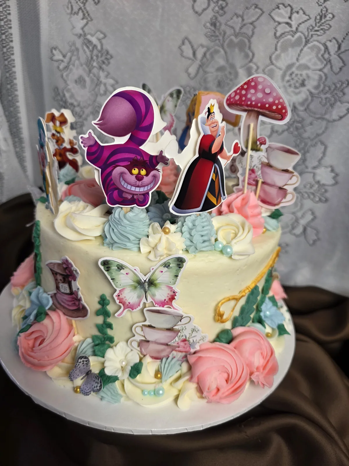 Alice in Wonderland birthday cake seen from the Cheshire Cat side, with the Queen of Hearts, toadstools, teacups, butterflies and pastel buttercream rosettes.