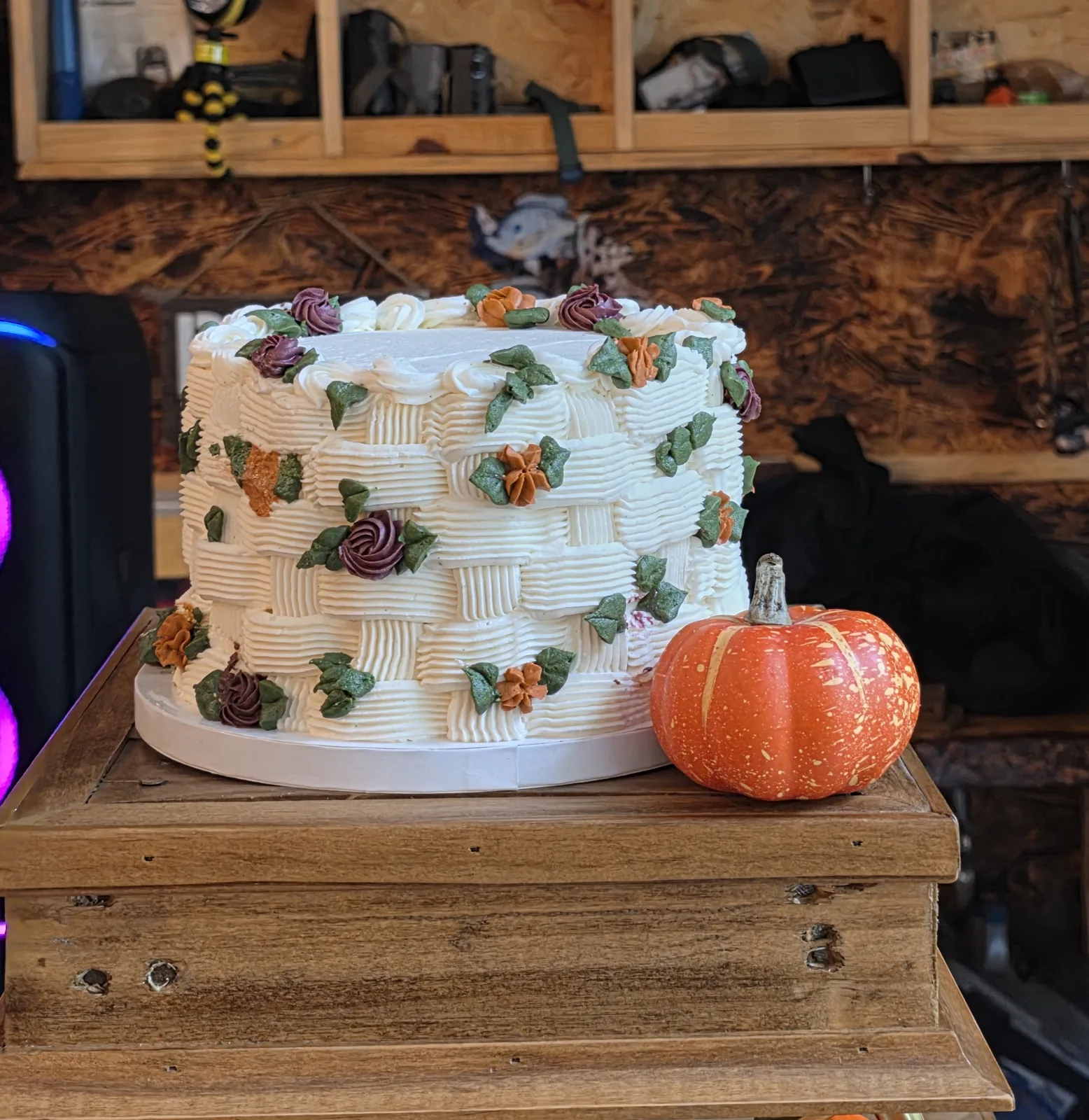 Small white wedding cake piped in a basket weave pattern with burgundy and orange buttercream flowers, beside a pumpkin on a rustic dessert table.