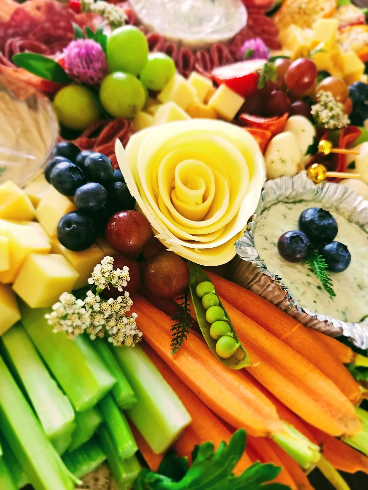 Close up of a charcuterie board with a cheese rose, carrots, celery, a snap pea, blueberries, cheese cubes and a herb dip.