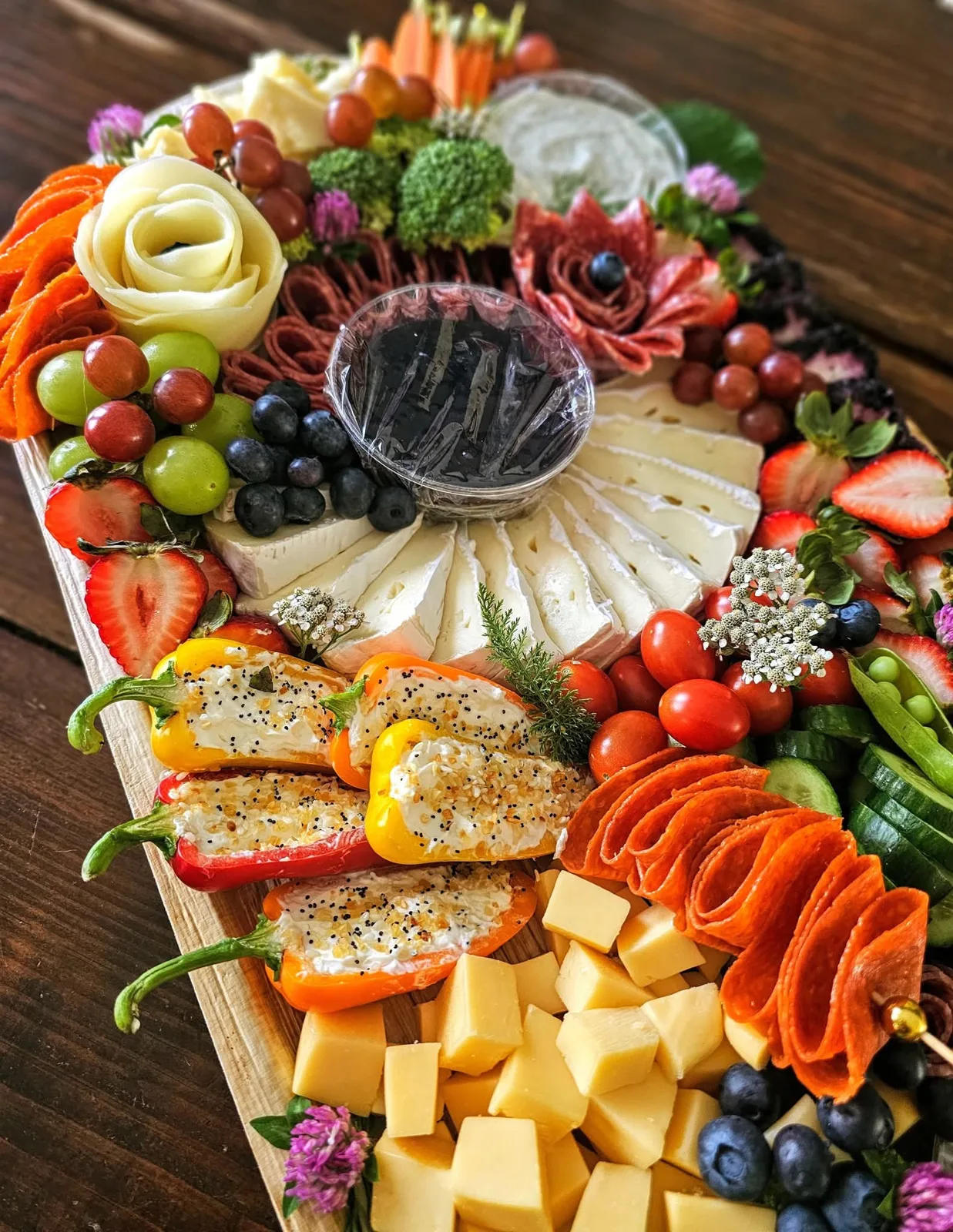 Charcuterie board with a cheese rose, a salami flower, brie wedges, stuffed mini peppers, folded pepperoni, cheese cubes, berries and a fig jam cup on a wooden board.