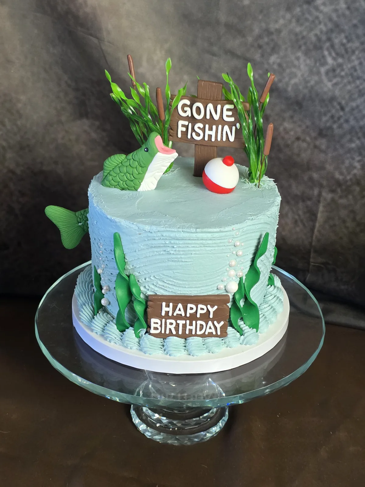 Gone Fishin themed birthday cake with a fondant bass, fishing bobber, cattails and a Happy Birthday sign on a glass stand.
