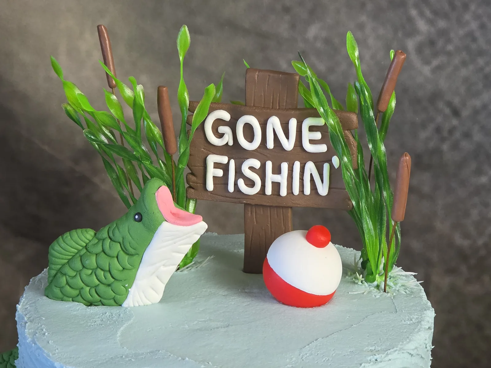 Close up of a Gone Fishin cake topper with a fondant bass, red and white bobber and cattails.