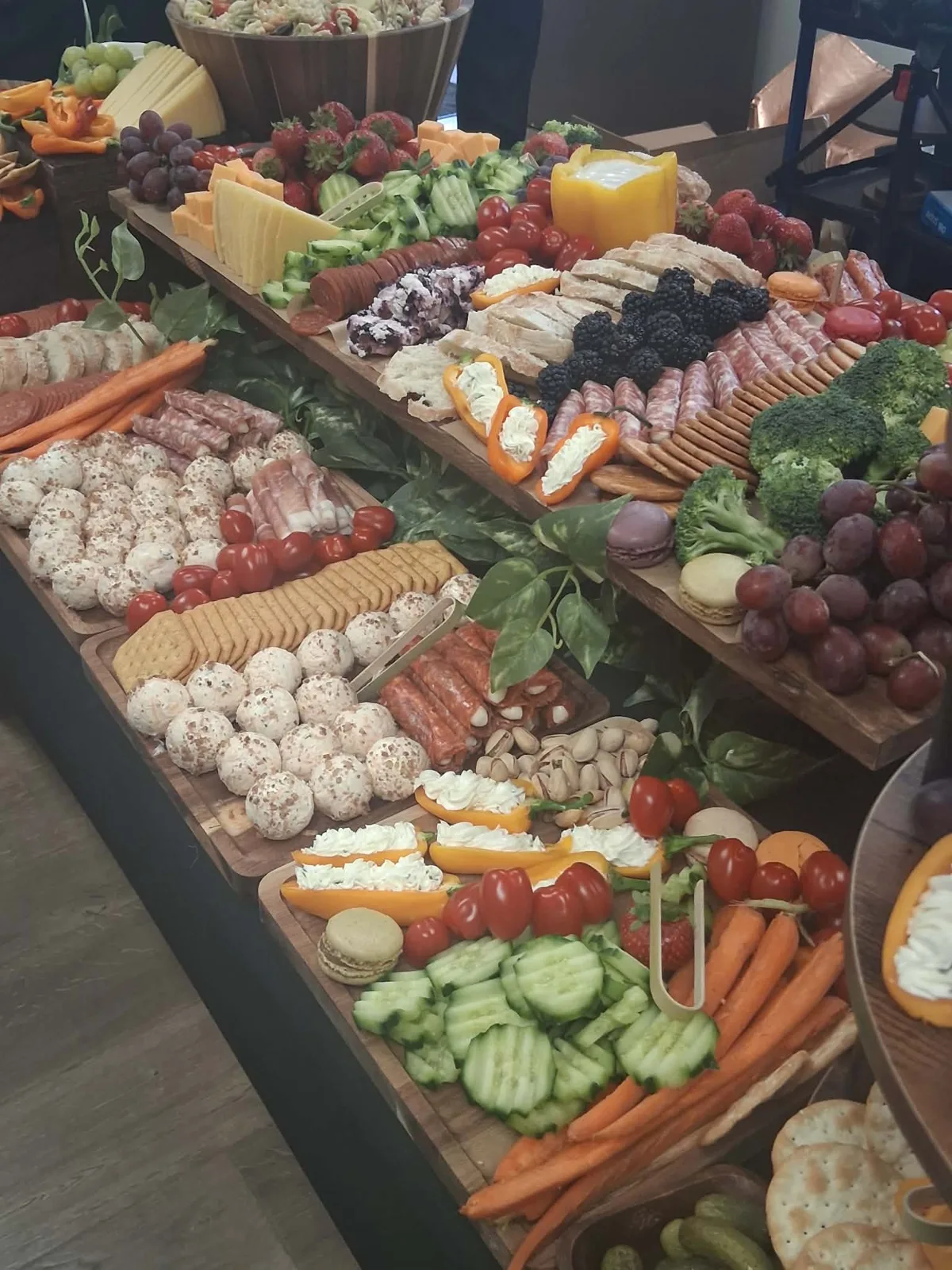 Two level grazing table with mini cheese balls, stuffed mini peppers, salami, crackers, broccoli, cucumbers, carrots, grapes and macarons.
