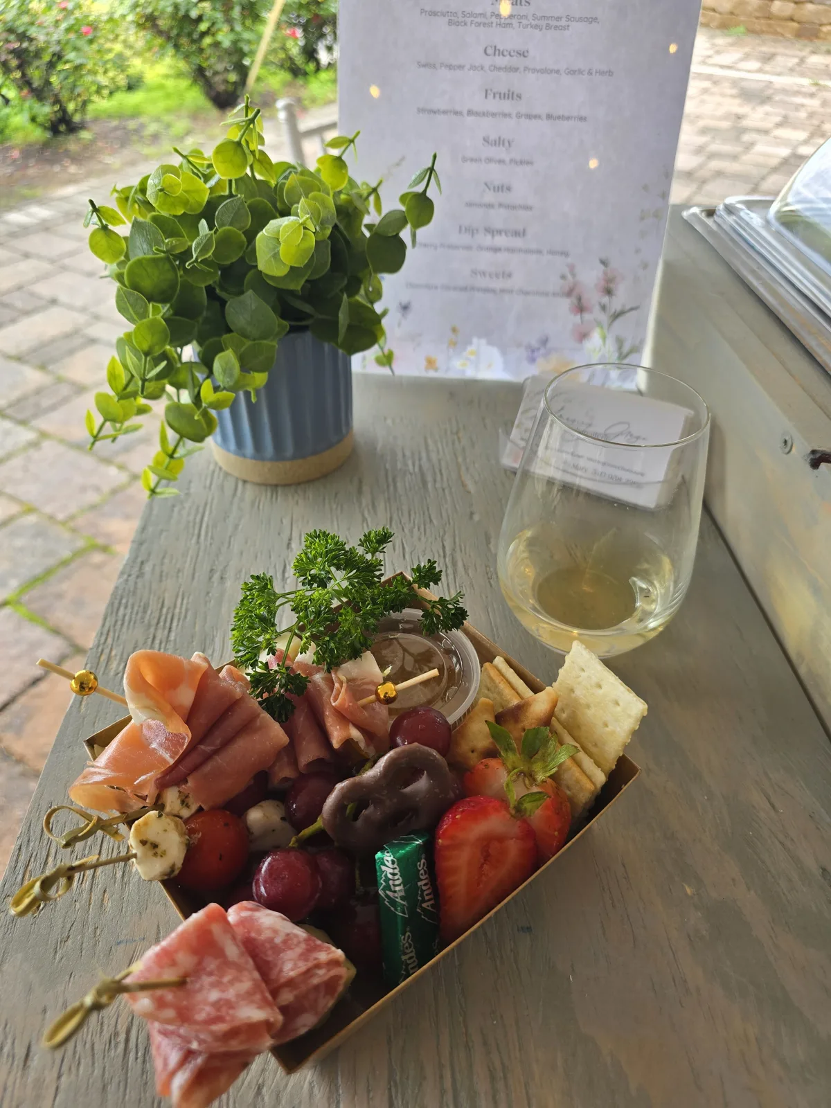Individual charcuterie box with prosciutto and salami skewers, mozzarella, grapes, a strawberry, crackers and a chocolate pretzel, served beside a glass of white wine.