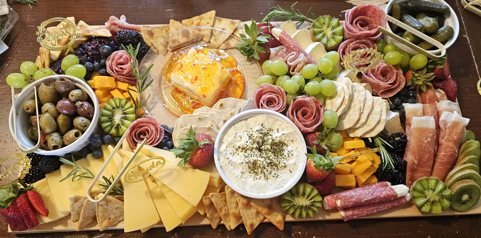 Large charcuterie board with salami roses, prosciutto, sliced cheeses, a pepper jelly and cream cheese centerpiece, a herb dip, olives, pickles, kiwi, grapes and crackers.