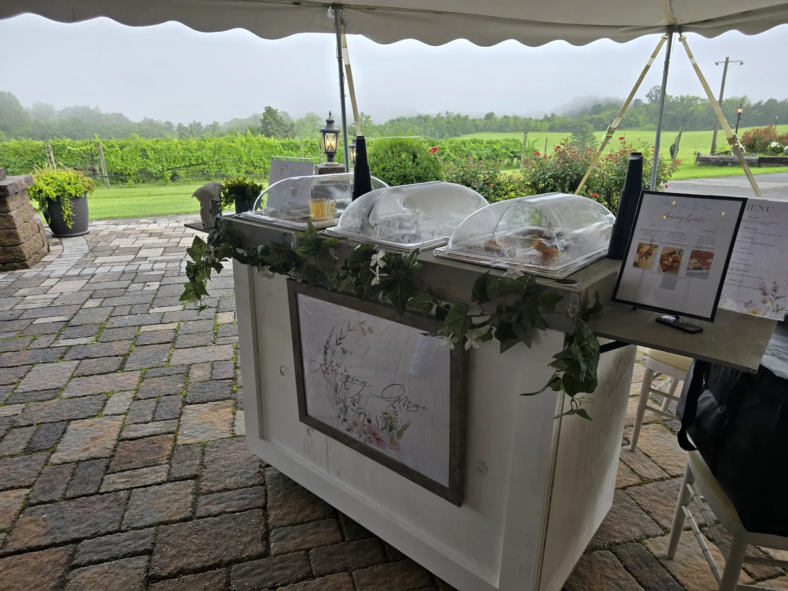 White mobile charcuterie cart trimmed with ivy, set up under a tent at a vineyard event with covered serving trays and a menu sign.