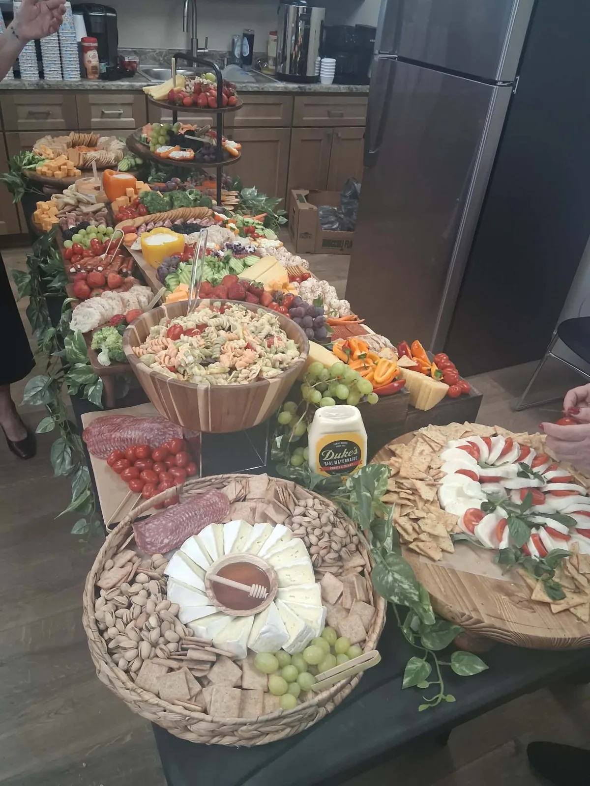 Long grazing table for an office event with a brie and honey basket, pistachios, caprese, pasta salad, tiered fruit stands and vegetable trays.