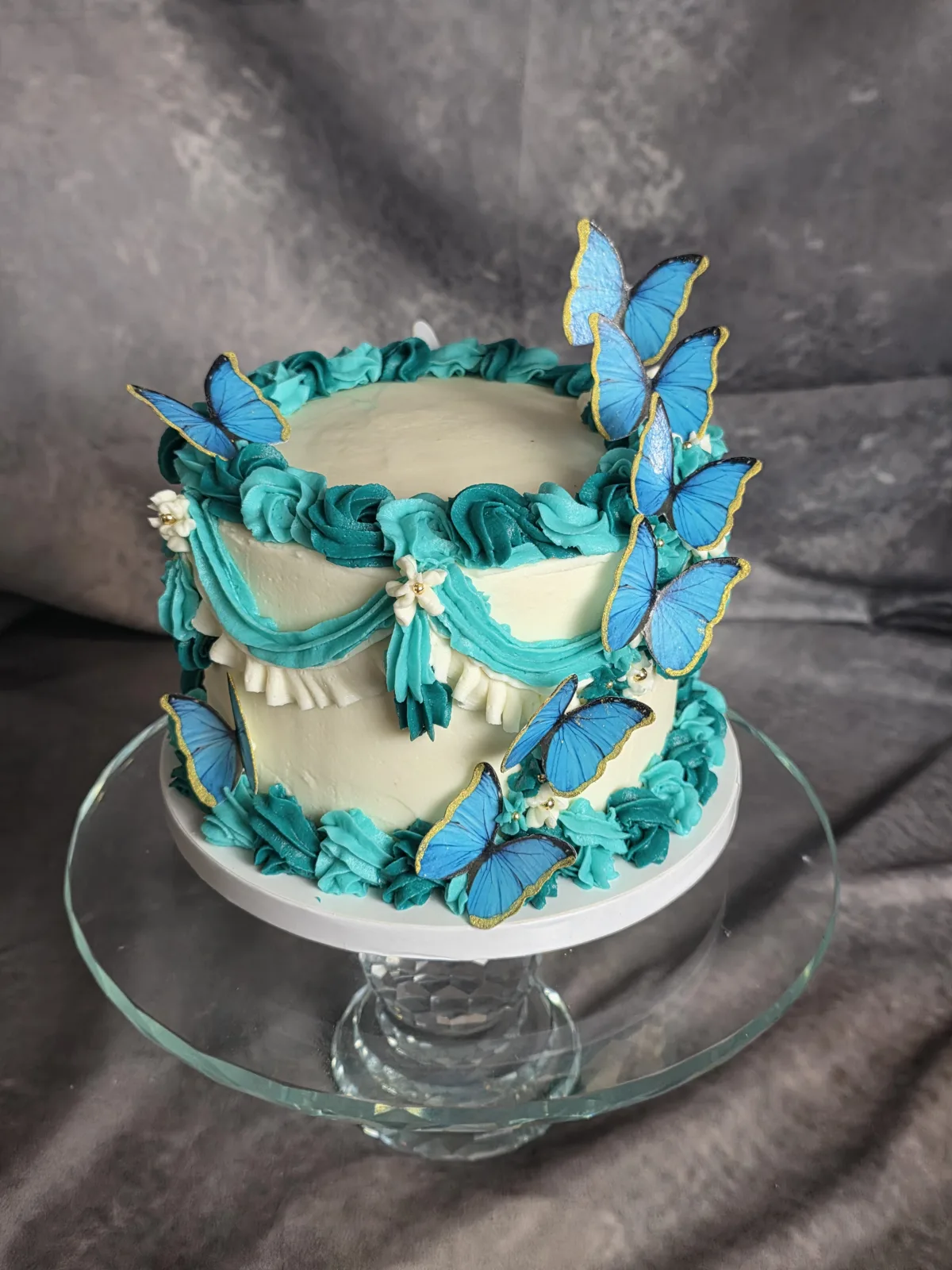 Vintage style buttercream birthday cake with teal swags and piping and blue butterflies on a glass cake stand.